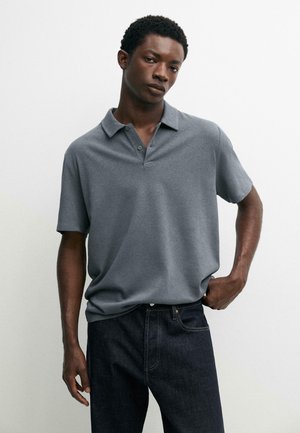 Giovane uomo dalla pelle scura che indossa una polo grigia a maniche corte e jeans scuri, in piedi davanti a uno sfondo bianco semplice.