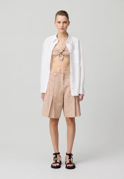 Top corto a rayas en beige y blanco con detalle de nudo, combinado con pantalones cortos beige con tablas. La modelo lleva sandalias negras con tiras. Camisa blanca de botones usada abierta.