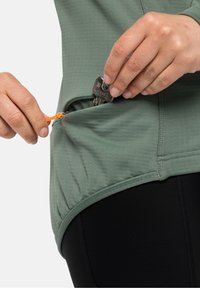 Top sportivo verde chiaro realizzato in tessuto testurizzato, con un tirante arancione e una tasca laterale per lo stoccaggio. Sotto sono visibili leggings neri.
