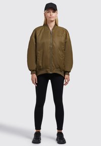 Olivovo zelená bomber bunda s rebrovanými manžetami a lemom, dvoma bočnými vrecami, zapínaním na zips a čiernym klobúkom. Nosená s čiernymi legínami a teniskami.
