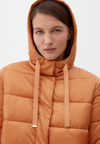 Kobieta o jasnej skórze i brązowych włosach nosi jasnobrązową pikowaną kurtkę z kapturem i długimi sznurkami na tle jasnym.