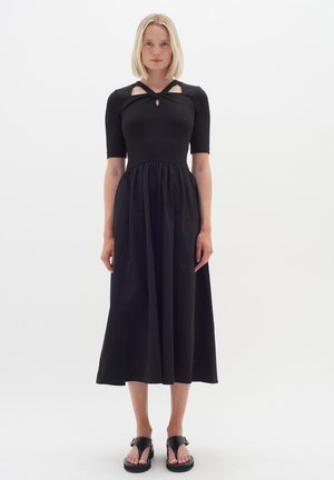 Abito maxi nero con top a coste e dettagli cut-out, maniche corte e gonna arricciata. La modella indossa sandali neri.