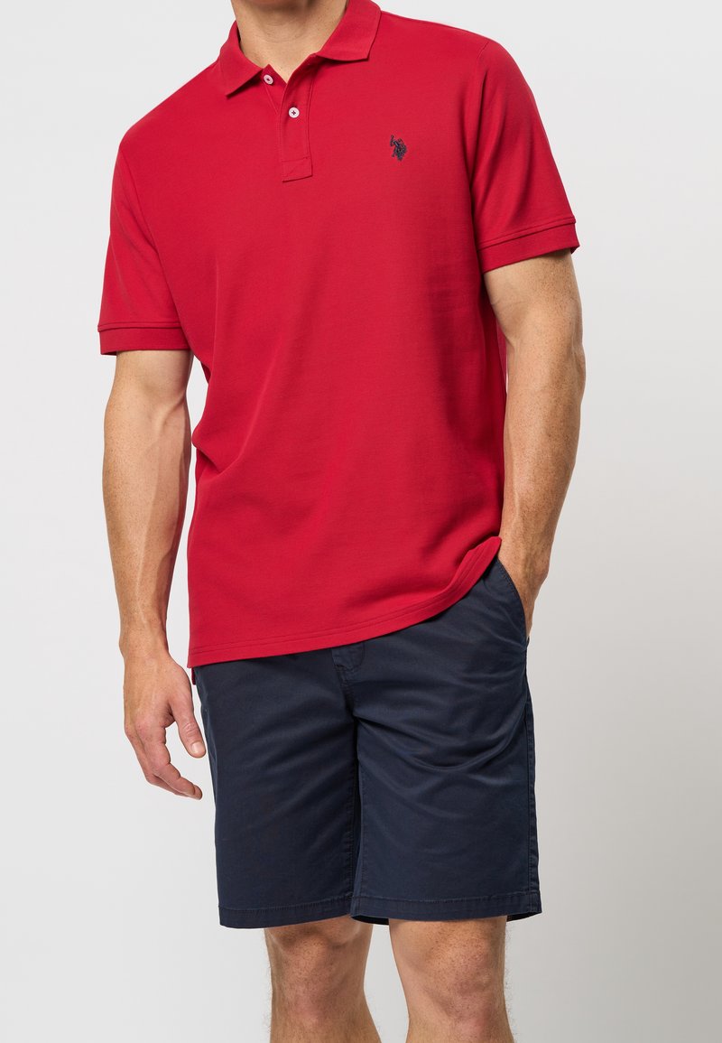 Polo rouge avec deux boutons, tissu texturé et logo bleu marine. Associé à un short bleu marine, avec une coupe décontractée et des poches.
