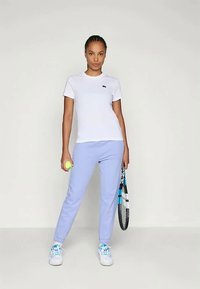 Camiseta blanca de manga corta con un pequeño logo azul, combinada con pantalones de chándal azul claro y zapatillas blancas, sosteniendo una pelota de tenis y una raqueta.