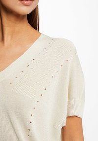 Kvinna iklädd en krämfärgad kortärmad stickad tröja med hålmönster och V-ringning, visad från halsen till midjan.