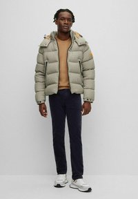 Giacca imbottita a quadri beige e marrone, con cappuccio, chiusura a zip e due tasche con zip. Indossata sopra un top beige e pantaloni blu navy.