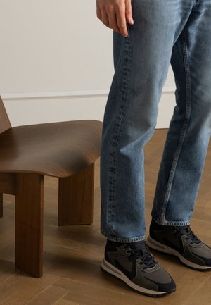 Jean en denim bleu avec une coupe droite, associé à des baskets noires et grises. À proximité, une chaise en bois courbée avec une finition lisse.