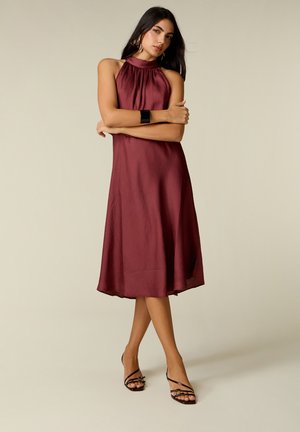 Vrouw met mouwloze bordeaux midi-jurk en zwarte sandalen met bandjes, staand met gekruiste armen tegen een effen achtergrond.