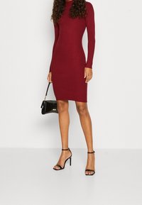 Burgunderrotes, geripptes Langarmkleid mit figurbetonter Silhouette. Kombiniert mit schwarzen, geschnürten High-Heels und einer kleinen, strukturierten Handtasche.