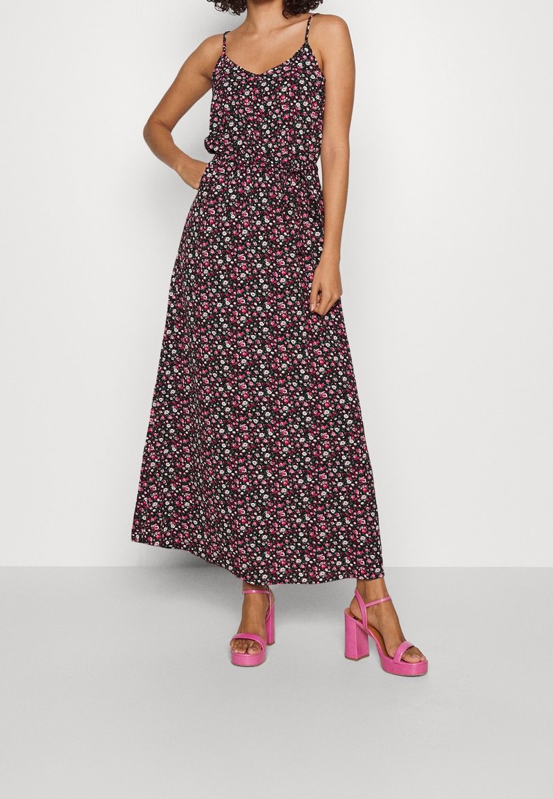 Robe maxi noire à motifs floraux avec des bretelles fines et une taille élastique. Présente un petit motif de fleurs en rose et blanc. Associée à des sandales à plateforme roses.