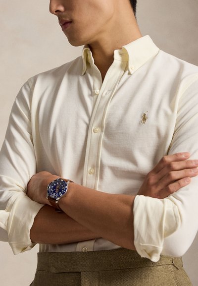 Mees, kes kannab kreemjat nööpidega särki rullitud varrukatega, käed rüpes ja nahast käepaelaga käekella, millel on sinine sihverplaat.