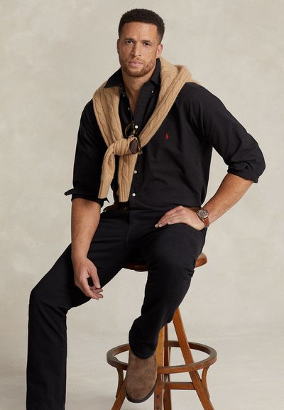 Man with short curly hair and freckles wears black shirt, tan sweater draped over shoulders, black pants, brown suede shoes, sitting on wooden stool.