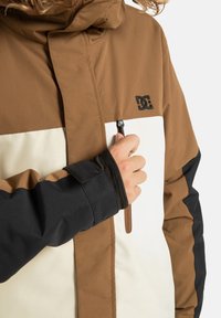 Giacca marrone e crema con dettagli neri, dotata di tasca con zip e polsini regolabili. Realizzata in tessuto liscio e duraturo.