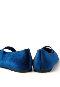 Des chaussures plates en velours bleu avec une texture douce, un bout arrondi et un détail de sangle élastique. La semelle est noire et légèrement courbée à l'arrière.