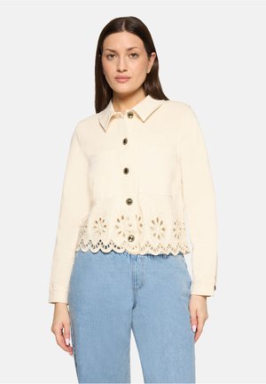 Frau trägt eine cremefarbene Jacke mit Knöpfen und einem gewellten Saum mit Blumen-Ausschnitten sowie hellblaue, hoch taillierte Jeans, steht vor einem schlichten Hintergrund.