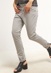 Pantalones slim-fit en gris claro, con cierre de botón y bolsillos frontales. Material suave y ligero. Llevados con tacones abiertos negros.