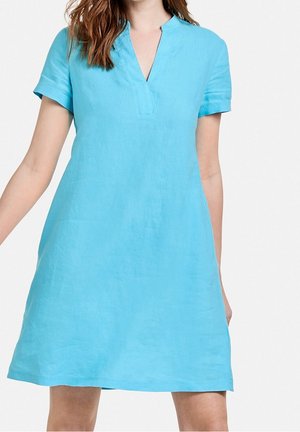 Vrouw met een turquoise jurk met korte mouwen, knielengte en een V-hals tegen een witte achtergrond.