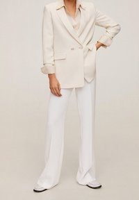 Dubbelzijdige crèmekleurige blazer met geknipte revers, gecombineerd met witte flared trousers. Zachte stof, nauwsluitende pasvorm en subtiel opgerolde mouwen.