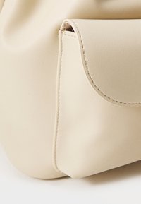 Sac à main en simili cuir beige avec un coin présentant une texture lisse, des coutures discrètes et une forme arrondie agrémentée d'un petit détail de poche à rabat.