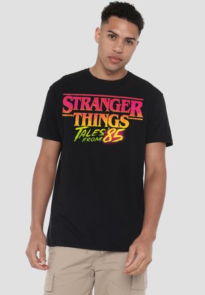Junger Mann trägt ein schwarzes T-Shirt mit buntem Schriftzug „Stranger Things Tales From 85“ und beigen Cargo-Shorts, steht auf grauem Hintergrund.