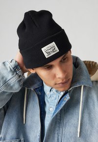 Zwarte geribbelde gebreide beanie met een rechthoekig wit label met zwarte logo-details. Draag het met een spijkerjack en een lichtblauw shirt eronder.