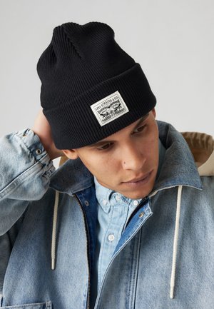 Schwarze gerippte Strickmütze mit einem rechteckigen weißen Patch und schwarzen Logo-Details. Getragen zu einer Jeansjacke und einem hellblauen Hemd darunter.