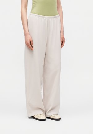 Άτομο φοράει φαρδιά ανοιχτό μπεζ παντελόνια με ελαστική μέση και παπούτσια κρεμ σε απλό λευκό φόντο.