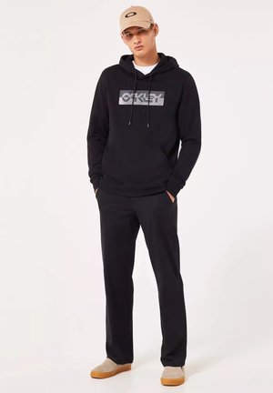 Giovane uomo che indossa una felpa con cappuccio nera Oakley, pantaloni neri, cappellino beige e scarpe beige, in piedi con le mani nelle tasche.