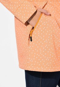 Chaqueta naranja repelente al agua con un patrón de gotas, bolsillo con cremallera y tirador naranja, y un ajuste entallado. Jeans de denim visibles por debajo.
