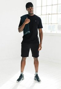 Mustar must pehme neel ja lühikesed püksid, must müts, hoides türkiissinist rätikut. Mustad spordijalatsid valgete aktsentidega. Erakordselt minimalistlik siseruum.