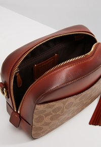 Ruskea nahkainen olkainlaukku, jossa on teksturoitu beige kangaspaneeli logokuvioinneilla, kullanvärinen vetoketju ja tumma kangasvuori sisällä.