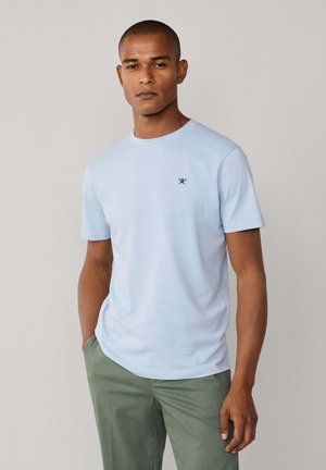T-shirt bleu clair à manches courtes, col rond, avec un petit emblème sombre sur la poitrine. Tissu en coton lisse, coupe décontractée.