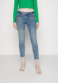 Vaqueros azules deslavados con un ajuste slim y largo recortado. Presentan bolsillos con cremallera y un cierre de botón. Combinados con una blusa verde y sandalias blancas.