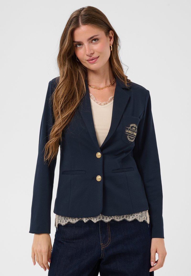 Femme aux longs cheveux bruns portant un blazer bleu foncé avec des boutons dorés et un top beige bordé de dentelle en dessous.