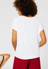 Maglietta bianca di cotone con maniche arrotolate e orlo arrotondato, indossata con pantaloni rossi texturizzati. Design minimalista, senza motivi o decorazioni.