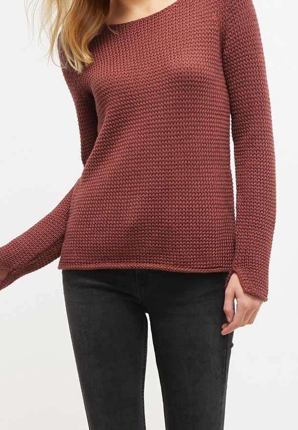 Femme portant un pull texturé à manches longues de couleur rouille et un pantalon noir, debout devant un fond clair uni.