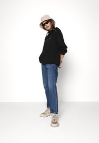 Czarna bluza z kapturem z kieszenią kangurką, niebieskie jeansy z wysokim stanem, beżowe i brązowe sneakersy, oraz różowy kapelusz w stylu wiaderka, na tle białym.