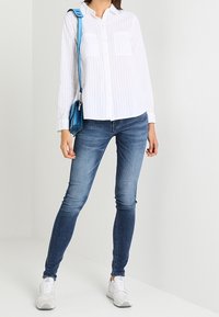 Kvinde iført hvid stribet skjorte med knap, slidte skinny jeans, hvide sneakers og med en blå crossbody taske over skulderen.