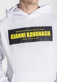 Biely mikina s veľkým čiernym obdĺžnikovým grafickým prvkom a žltým textom "DBAJTE NA REPLIKY GIANNI KAVANAGH, VYROBENÉ PRE VÍŤAZOV."