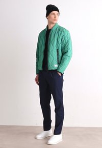 Quilted green bomber jacket with a stand collar, zippered pockets, paired with navy pants and white sneakers. Black beanie completes the look.