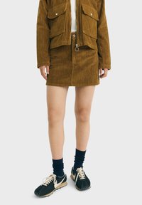 Braune Kordjacke mit zwei Fronttaschen, kombiniert mit einem passenden braunen Kordrock. Schwarze Sneaker mit weißen Akzenten und marineblauen Socken.