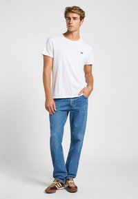 Camiseta de algodón blanca con el logo negro de "Lee" en el lado izquierdo del pecho, combinada con jeans de mezclilla azul y zapatillas marrones, rojas y amarillas.