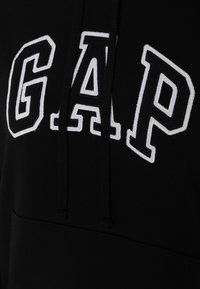Sudadera negra con un destacado logo "GAP" bordado en blanco. Incluye capucha con cordones y un bolsillo delantero. Textura lisa.