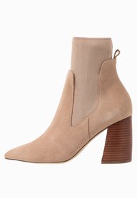 Beżowy zamszowy botek sięgający do kostki z tkaną cholewką przypominającą skarpetkę, spiczastym noskiem i żebrowanym drewnianym obcasem. Gładka tekstura na całej powierzchni z elastycznymi bocznymi panelami.
