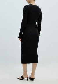 Zwarte ribgebreide jurk met lange mouwen, getailleerde silhoutte en een lengte tot halverwege de kuit. Ggedragen met elegante zwarte hoge hakken.