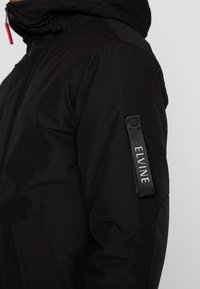 Czarna pikowana kurtka z zamkiem błyskawicznym z przodu i kapturem, wykonana z fakturowanego materiału, z bocznymi kieszeniami oraz oznakowaną metką na rękawie.