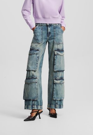 Personne portant un jean cargo large délavé bleu avec plusieurs grandes poches et des chaussures à talons noires, assorti à un sweat-shirt lavande.