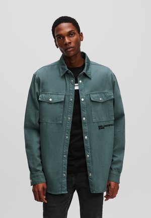 Teal denim shirt with two chest pockets, silver hardware, and embroidered branding on the left side, layered over a black shirt.