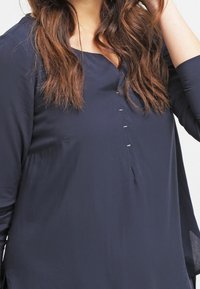 Blusa blu navy in tessuto leggero, con scollatura rotonda, dettaglio dei bottoni lungo la parte frontale e maniche lunghe. Texture liscia.