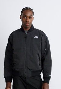 Homme avec des cheveux tressés portant une veste bomber à fermeture éclair noire de North Face sur un pantalon sombre, faisant face à un fond blanc uni.